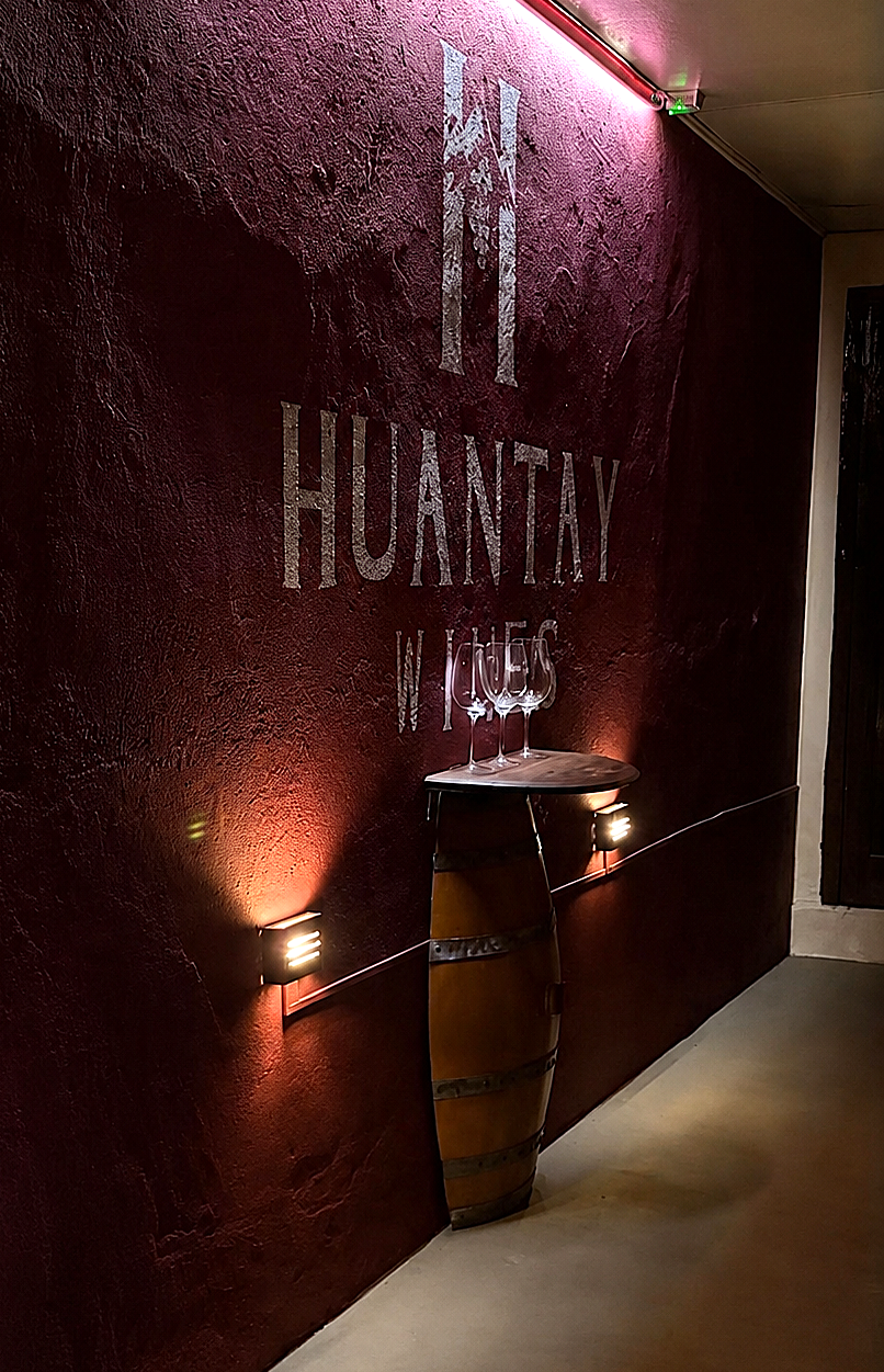 Pasillo interior de Huantay Wines con pared bordó iluminada, barrica y copas